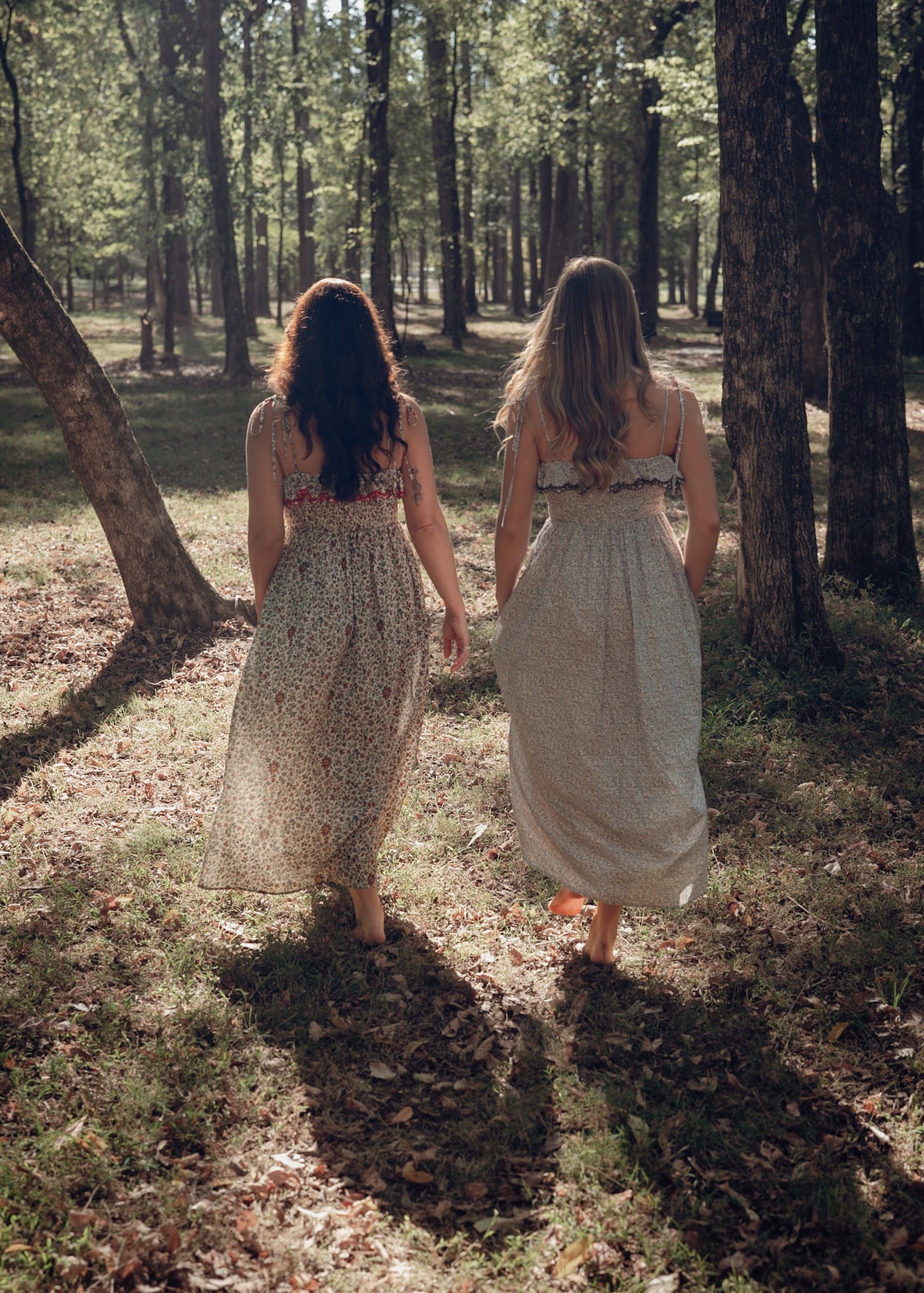 Two models walking barefoot through the woods in the Avaire Collective Celeste Dress, one in Red and one in Blue, seen from behind — homepage hero
