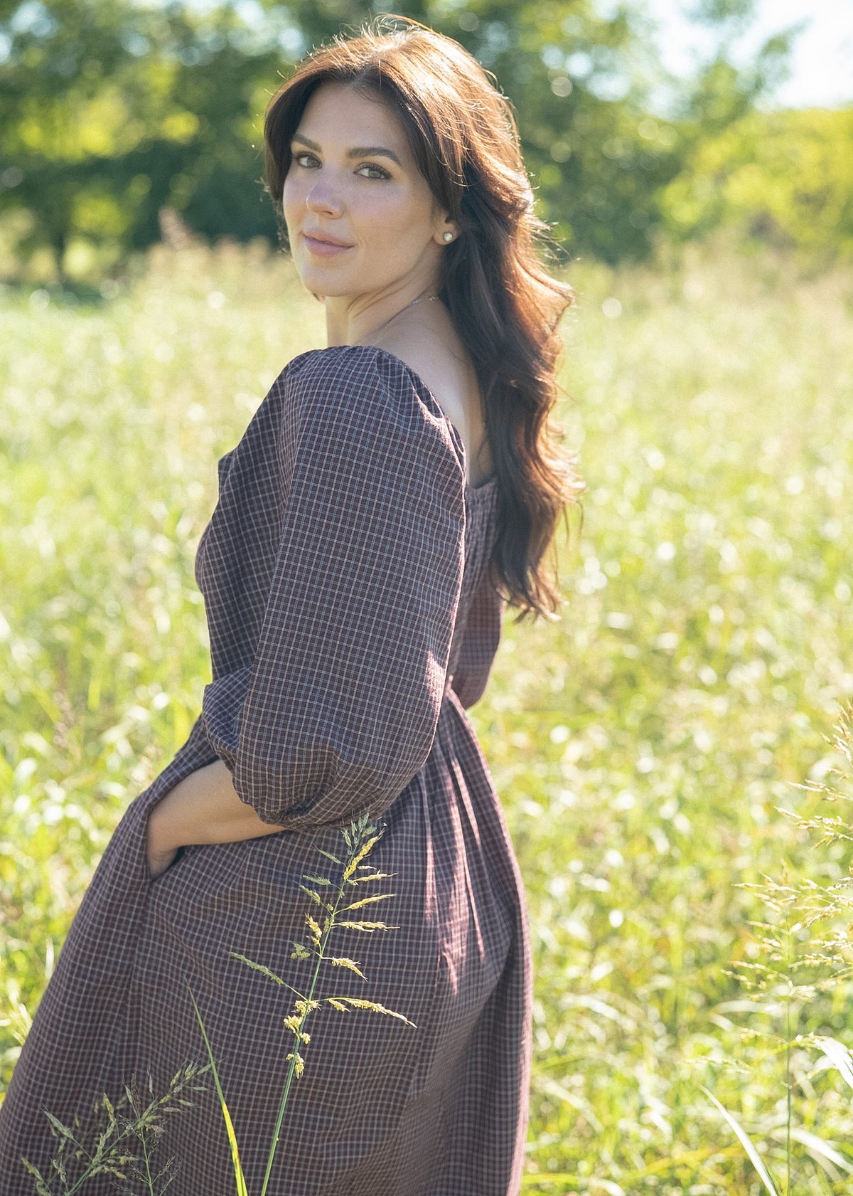 Woman in the Avaire Collective Vienna Dress, a plum plaid cotton dress with balloon sleeves, looking back over her shoulder in a sunlit meadow